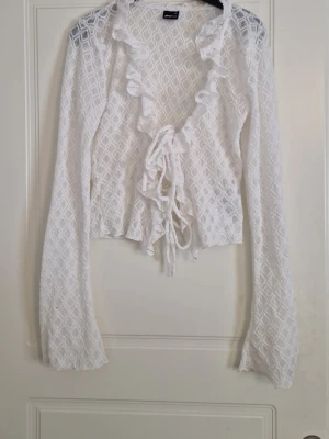 Vit spetsig bolerotopp med volang - Superfin vit bolerotopp från Gina tricot i spetsliknande material. Toppen har långa ärmar, volangkant längs urringningen och knyt framtill. Perfekt för att lyfta en outfit med sin romantiska look och genombrutna mönster.