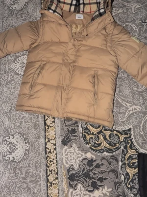 Burberry jacka - Snygg beige pufferjacka med dragkedja och knappar framtill. Jackan är gjord av premium material som hållerr en varm och skön i vintern. Man kan även dra av armarna och göra den till en väst för extra style och för varma sommar dagar.
