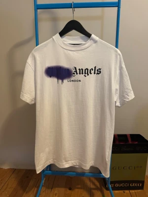 Vit Palm Angels t-shirt med tryck - Säljer en vit t-shirt från Palm Angels med svart text och lila spray paint-effekt på bröstet. Modellen är klassisk med rund hals och korta ärmar. Perfekt för dig som gillar streetwear och vill sticka ut lite extra. Storlek M