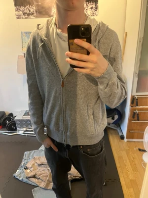 Grå zip-hoodie Kashmir - Säljer en grå hoodie med dragkedja och huva. Modellen har ribbade muddar, fickor framtill och är tillverkad i kashmir. Perfekt för en avslappnad och stilren look.