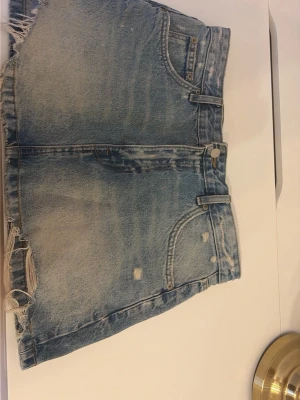 Blå jeanskjol med hål  - Snygg blå denimkjol med mellanmidja och fransig kant nertill. Kjolen har slitna detaljer och klassisk femficksdesign. Perfekta för sommardagar och ger en avslappnad vibe.