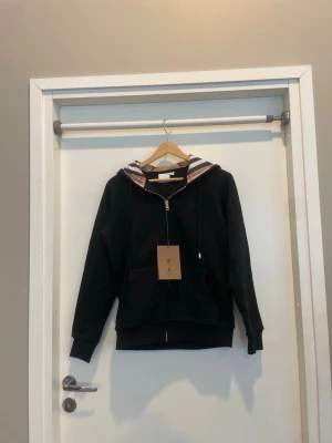 Svart hoodie med Burberry-detaljer - Svart hoodie med dragkedja från Burberry. Klassisk huva med det ikoniska rutmönstret på insidan och snörning. Mjuk bomullskänsla, fickor framtill och ribbade muddar. Perfekt för en chill och stilren look.