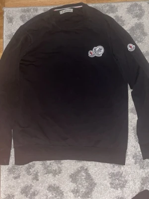 Svart sweatshirt från Moncler - Svart sweatshirt från Moncler med rund halsringning och ribbade muddar. Tröjan har Moncler-logga på bröstet och ärmen, vilket ger en exklusiv känsla. Perfekt för dig som gillar stilrena och klassiska plagg med premiumkänsla.