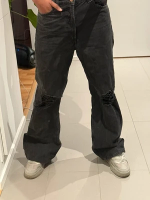 Svarta jeans med hål - Säljer ett par svarta baggy jeans med slitna hål över knäna. Jeansen har en lös passform och raka ben, perfekt för en avslappnad streetstil. Klassisk och knappgylf. Materialet är denim i bomull.