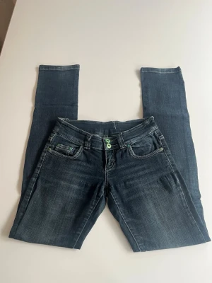 Mörkblå raka jenas  - Snygga mörkblå raka jeans med kontrastsömmar och dekorativ broderad bakficka. Jeansen har låg midja, dubbla gröna knappar och klassisk femficksdesign. Tillverkade i stretchigt denim för skön passform.