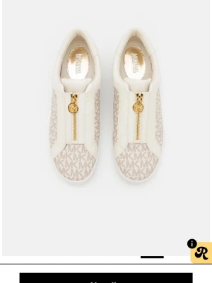 Michael Kors vita sneakers med dragkedja - Snygga vita sneakers från Michael Kors med beige MK-monogram på sidorna och guldfärgad dragkedja framtill. Skorna har en låg profil och är tillverkade i en mix av textil och konstläder. Perfekta för dig som gillar stilrena detaljer och logomönster.