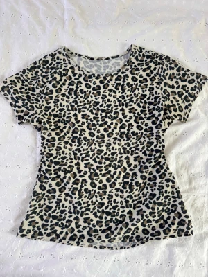 Leopardmönstrad t-shirt i bomull - Cool t-shirt med leopardmönster i svart och brunt på beige botten. Klassisk rund halsringning och korta ärmar. Perfekt för dig som vill sticka ut med en trendig look. Skön och mjuk känsla tack vare bomullsmaterialet.