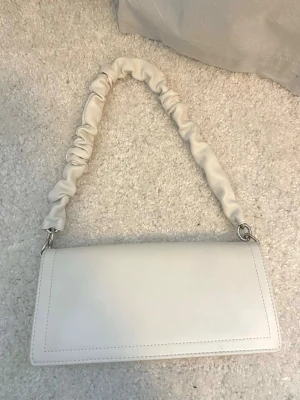 Vit axelremsväska Gina Tricot - Trendig vit handväska (bärs över axeln) i rektangulär form med en unik rynkad axelrem. Väskan har magnetlås och silverfärgade metalldetaljer. Tillverkad i slätt konstläder som ger en stilren look. 