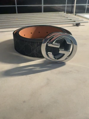 Svart Gucci skärp med stor silverlogga - Snyggt svart skärp från Gucci med klassiskt GG-mönster och stor, rund silverfärgad logga som spänne. Insidan är beige och bältet är gjort i äkta skinn. Perfekt accessoar för att lyfta vilken outfit som helst.