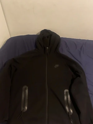 Svart Nike Tech - Svart Nike Tech från Nike med dragkedja hela vägen, huva och två fickor med dragkedja. Klassisk Nike-logga på bröstet. Perfekt för träning eller chill. Mjuk och bekväm bomullsblandning.