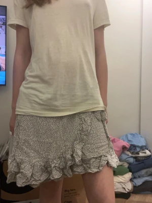 Grå omlottkjol med volanger - Supersöt grå kort omlottkjol med små vita blommor och volangkant. Kjolen har knyt i sidan och är gjord i ett lätt och luftigt material. Storlek S köpt på Non marknad i Spanien. 