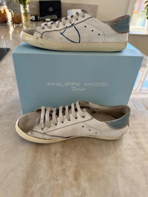 Philippe Model vita sneakers med blå detalj - Snygga vita sneakers från Philippe Model Junior med blå häl och grå detaljer. Skorna har klassisk låg profil, vita snören och en ikonisk blå logga på sidan. Tillverkade i läder med slitstark gummisula. Det ingår även en låda 📦🔥