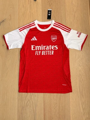 Arsenal matchtröja röd/vit Adidas - Snygg Arsenal matchtröja från Adidas i rött och vitt med klubbmärke och sponsortryck på bröstet. Tröjan har vita ärmar, röda detaljer och klassiska Adidas-ränder på axlarna. Tillverkad i lätt och ventilerande funktionsmaterial, perfekt för fotbollsträning.