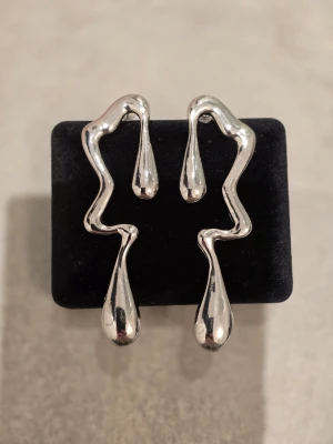 Silvriga statement örhängen i droppform - Unika silvriga örhängen med vågig, abstrakt form och stora droppdetaljer. De är iögonfallande och har en modern design som verkligen sticker ut. Perfekta för dig som gillar att bära något annorlunda och trendigt.