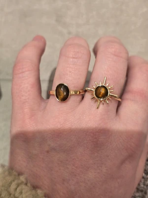 Två guldfärgade ringar med tigeröga - Säljer två snygga guldfärgade ringar med bruna tigeröga-stenar. Den ena ringen har en enkel oval sten, medan den andra har en solinspirerad design med strålar runt stenen. Perfekta för dig som gillar boho och unika detaljer. Justerbara storlekar