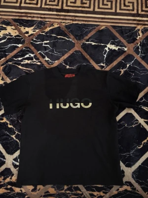 Svart HUGO t-shirt med logga - Svart t-shirt från HUGO med stor logga i vitt och grått framtill. Klassisk rund halsringning och korta ärmar. Perfekt för en stilren och enkel look. Tillverkad i mjuk bomull för skön komfort.