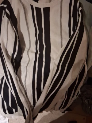 Fin GANT vit/svart stickad tröja med svarta ränder - Snygg stickad tröja i beige med svarta och gråa vertikala ränder. Tröjan har rund halsringning och långa ärmar. Perfekt för dig som gillar en stilren och modern look med lite edge.