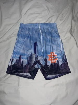 Blå Eric Emanuel shorts New York - Säljer ett par blå meshshorts med tryck av en stadssilhuett och en orange logga på ena benet. Shortsen har elastisk midja med snörning och är perfekta för en avslappnad stil. Materialet är lätt och andas, vilket gör dem sköna under varma dagar.