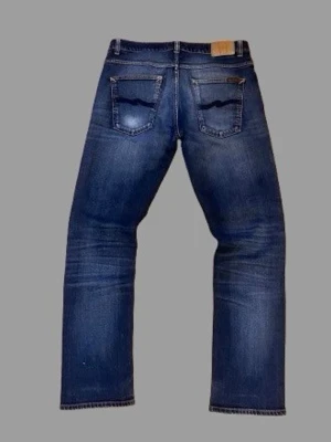 raka nudie jeans - Snygga blå jeans med raka ben och slitna detaljer. Jeansen har klassiska fem fickor, ljusare tvättade partier och råa kanter vid bensluten. Perfekta för en avslappnad och trendig look.