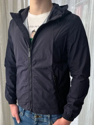 Mörkblå Censured vindjacka, storlek S - Marinblå/mörkblå vindjacka (windbreaker) från Censured i stilren och modern design. En lätt och praktisk jacka som passar perfekt för vår, sommar och tidig höst.  Jackan är använd en del men fortfarande i mycket bra skick. Inga defekter – fungerar helt som den ska med dragkedjor och detaljer i gott skick.  Storlek S. Mått: • Axelbredd: ca 45 cm • Längd: ca 65 cm (utan luva)  Hör gärna av dig vid frågor eller om du önskar fler bilder!