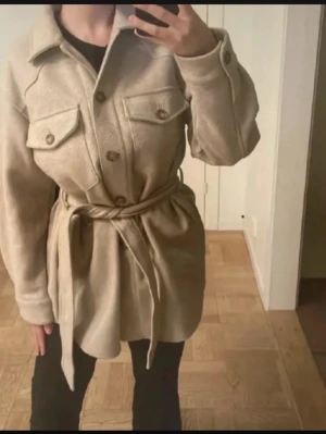 Beige overshirt med knytskärp - Stilren beige overshirt med stora bröstfickor, knappar framtill och ett matchande knytskärp i midjan. Overshirten har klassisk krage och är tillverkad i ett mjukt, ull-liknande material. Perfekt för lager på lager och ger en avslappnad men trendig look.