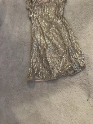 Glittrig silver kortklänning - Säljer en glittrig kortklänning i silver med paljetter över hela klänningen. Klänningen har en figurnära passform och är ärmlös, perfekt för dig som vill sticka ut. Ingen synlig logga eller märke.