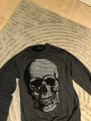 Mörkgrå tröja från Philipp Plein -  mörkgrå tröja från Philipp Plein med stort tryck framtill och logotypen tryckt längs ärmarna. Tröjan har rund hals och långa ärmar. Perfekt för dig som gillar exklusiva märken. Den här tröjan passar även för storlek L 