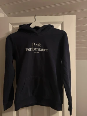 Mörkblå hoodie från Peak Performance - Snygg mörkblå hoodie från Peak Performance med vit broderad logga på bröstet. Klassisk ficka framtill och huva. Tillverkad i mjukt och bekvämt material som passar perfekt till chill dagar. Enkel och stilren design som funkar till det mesta.
