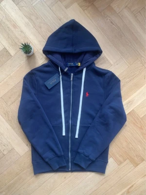 Marinblå hoodie från Polo Ralph Lauren - Snygg marinblå hoodie med dragkedja från Polo Ralph Lauren. Har vit dragsko i huvan och klassisk röd logga broderad på bröstet. Mjuk bomullsblandning och kängurufickor framtill. Perfekt för en chill och stilren look.