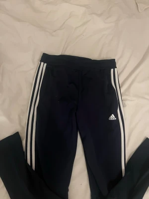 Marinblå Adidas träningsbyxor - Säljer ett par marinblå träningsbyxor från Adidas med de klassiska vita ränderna längs sidorna och Adidas-logga på vänster ben. !!Om du köper välj gärna att hämta vid upphämtningsställe istället för hemleverans då det blir enklare för mig att posta på det viset!!