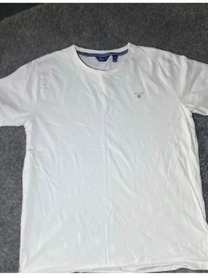 Vit T-shirt från Gant i bomull - Klassisk vit T-shirt från Gant med rund halsringning och diskret logga broderad på bröstet. Tillverkad i mjuk bomull och har en normal passform. Perfekt basplagg till garderoben.