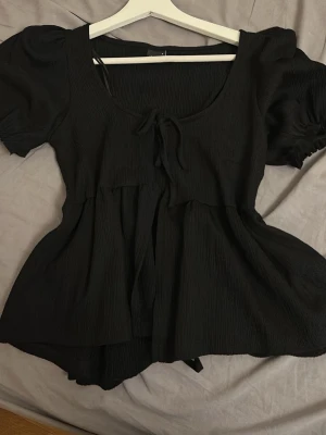 Svart blus med puffärm och knyt - Säljer en svart blus med puffärmar och knyt framtill. Blusen har en rundad urringning och ett flowy fall nedtill. Materialet känns lätt och har en diskret struktur. Perfekt för dig som gillar en söt och trendig look.