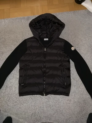Moncler cardigan äkta  - Snygg svart pufferjacka från Moncler med huva och dragkedja framtill. Jackan har quiltad kropp i glansigt material och svarta ärmar i mjukare tyg. Moncler-logga på vänster ärm. Perfekt för kalla dagar och riktigt trendig stil. Storlek L men passar som M knappt använd helt ny 