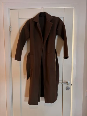 Brun lång kappa med skärp - Stilren brun kappa i lång modell med klassisk krage och stora fickor framtill. Kappan har ett matchande knytbälte i midjan och raka ärmar. Perfekt för dig som vill ha en enkel men snygg look under kallare dagar.