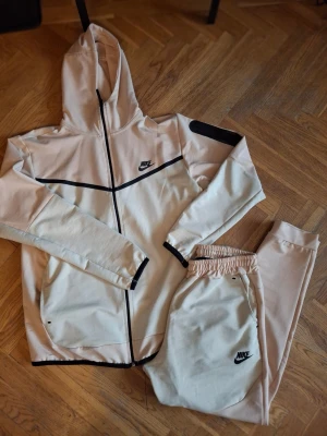  Nike set - Snyggt från Nike med matchande hoodie och byxor. Setet har svarta detaljer, dragkedja på hoodien och elastisk midja på byxorna. Båda delarna har Nike-logga tryckt framtill. Färgen är aprikos/beige. Använd en gång.