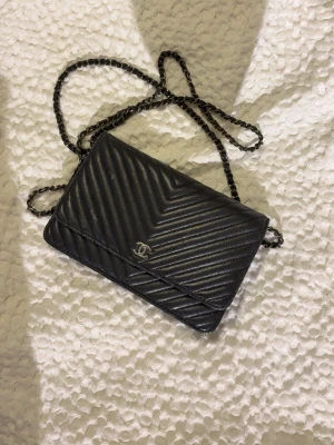 Svart kuvertväska i läder - Snygg svart kuvertväska från Chanel i quiltat läder med klassiskt V-mönster. Väskan har en silverfärgad kedjerem och ikonisk CC-logga framtill. Insidan är fodrad i vinrött tyg och har dragkedjefack. Perfekt för att bära det viktigaste med stil.