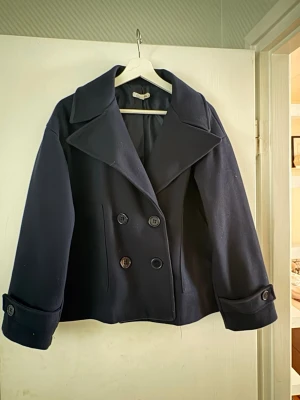 Mörkblå dubbelknäppt ullrock - Snygg mörkblå rock i ullmix med bred krage och dubbelknäppning framtill. Storlek xs/s  Kortare modell med dekorativa knappar vid ärmsluten och två sidofickor. Perfekt för dig som gillar klassisk men trendig stil.