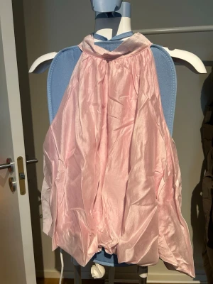 Ljusrosa halterneck topp - Säljer en ljusrosa halterneck topp i glansigt satinliknande material. Toppen är ärmlös och har en lös passform med hög halsringning. Perfekt för dig som vill ha en chic och trendig look. Knyts i nacken. 