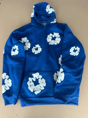 Blå hoodie med vita moln - Säljer en blå hoodie med huva och vita molnliknande mönster över hela plagget. Hoodien har ribbade muddar vid ärmslut och nederkant samt en klassisk känguruficka framtill. Perfekt för dig som vill sticka ut med en färgglad och unik stil.