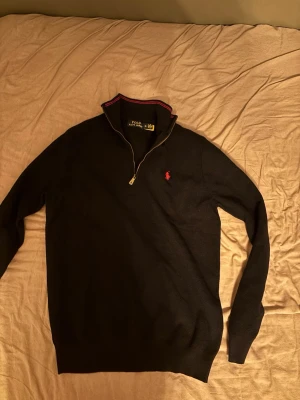 Svart tröja med dragkedja från Polo Ralph Lauren - Klassisk svart tröja från Polo Ralph Lauren med halv dragkedja och röd broderad logga på bröstet. Tröjan har ribbade muddar, hög krage med röd detalj och är perfekt för lager på lager. Materialet känns mjukt och bekvämt.