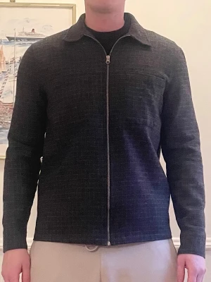 Grå overshirt från A Day's March - Stilren grå overshirt från A Day's March med diskret rutmönster. Dragkedja framtill, klassisk krage och två bröstfickor. Tillverkad i ett mjukt, vävt material som ger en avslappnad men ändå uppklädd look. Perfekt att bära över en t-shirt. Den är rätt varm så perfekt för tidig vår! Nypris 2299kr