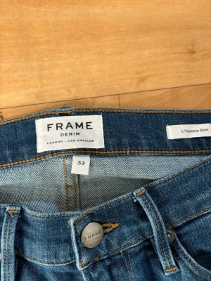 Frame Denim - Säljer ett par blå slim fit jeans från Frame Denim, modell L'Homme Slim. Klassisk femficksmodell med snygga slitningar och kontrastsömmar. Jeansen har knappgylf och är tillverkade i mjukt denimtyg som ger en skön passform.