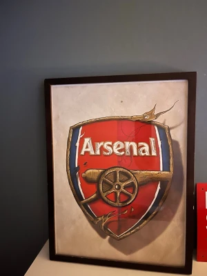 Arsenal tavla med träram - Snygg tavla med Arsenal-logga i rött, vitt och blått. Motivet är tryckt på papper och inramat i en mörkbrun träram. Perfekt för dig som gillar fotboll och vill ha något unikt på väggen.