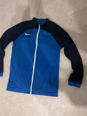 Blå träningsjacka från Nike - Snygg blå träningsjacka från Nike med svarta ärmar och vit dragkedja. Jackan har två fickor med dragkedja och den klassiska Nike-loggan på bröstet. Perfekt för träning eller uppvärmning.