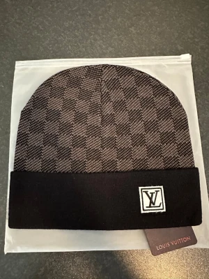 Louiss Vuitton svart och grå mössa - Snygg svart och grå mössa från Louiss Vuitton med klassiskt rutmönster och vikt kant. L.V-logga framtill på en vit patch. Tillverkad i mjukt material som känns lyxigt och bekvämt. Perfekt accessoar för dig som gillar exklusiv streetstyle tiktok.Följ marknads.closet