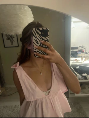 Ljusrosa ärmlös linnetopp med rosetter - Supersöt ljusrosa topp med djup v-ringning och breda axelband som knyts i rosetter. Modellen är ärmlös och har en luftig, voluminös passform. Perfekt för dig som gillar en feminin och trendig stil. Använd 2 gånger