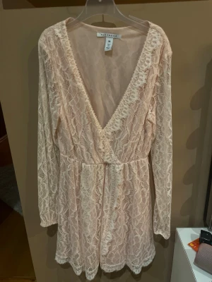 Ljusrosa spetsklänning  - Söt ljusrosa kortklänning med v-ringning och långärmad design. Klänningen är täckt av ett vackert spetsmönster och har omlott framtill. Perfekt för dig som gillar romantisk och feminin stil.