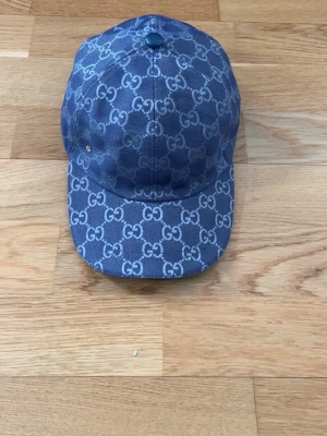 Blå keps från Gucci med GG-mönster - Snygg blå keps från Gucci med klassiskt GG-mönster över hela kepsen. Kepsen har böjd skärm och justerbar rem baktill. Materialet är denim-liknande tyg och insidan är ljus med Gucci-taggar. Perfekt för dig som vill ha en trendig accessoar.