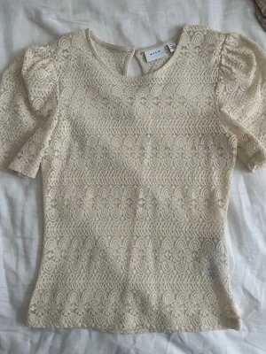 Beige spetsblus från Vila - Supersöt beige spetsblus från Vila med puffärmar och rund halsringning. Blusen har fina spetsdetaljer över hela plagget och en liten knapp i nacken. 