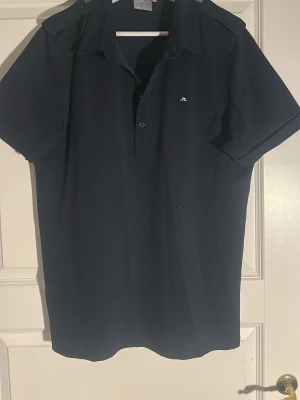 Marinblå pikétröja från J.Lindeberg - Snygg marinblå pikétröja från J.Lindeberg med korta ärmar och klassisk krage. Tröjan har knappar framtill och axelklaffar som ger en sportig touch. Tillverkad i mjuk bomull för skön känsla. Liten logga broderad på bröstet. Storleken är som L 